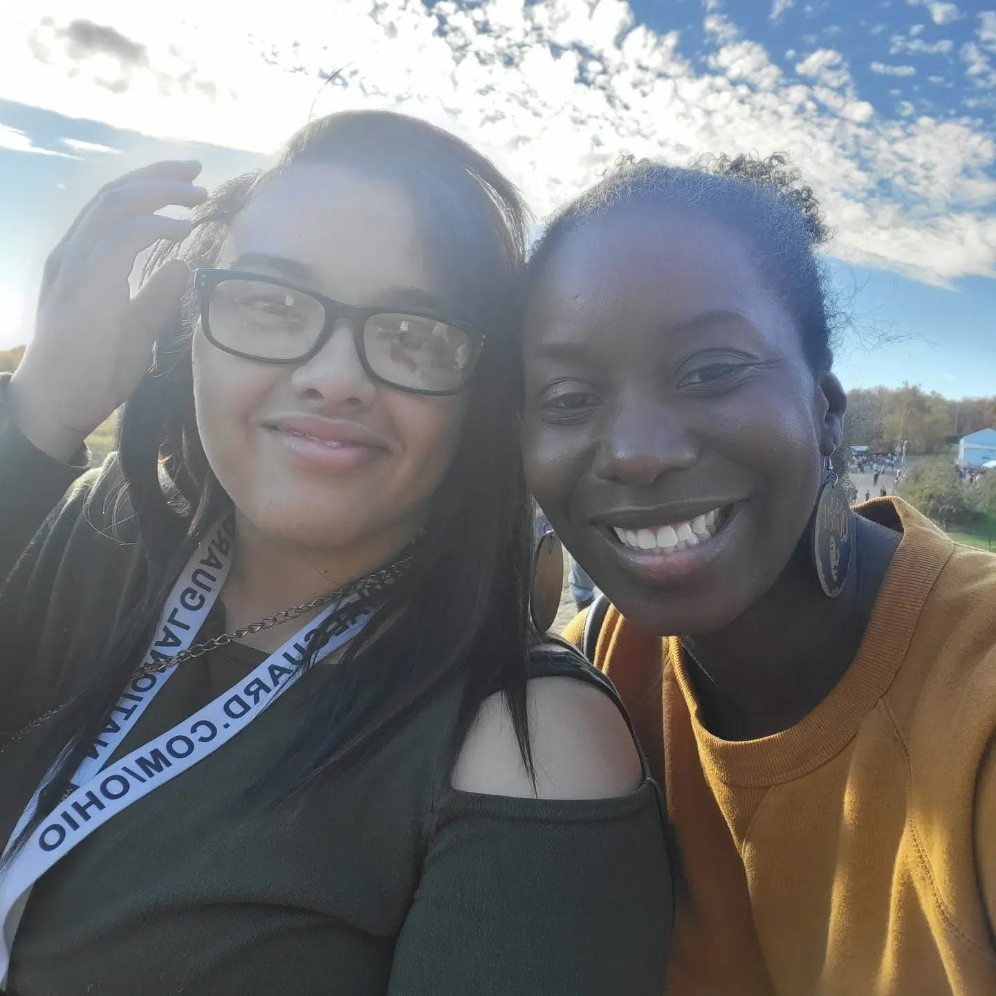 A Big Sister and Little Sister posing outdoors against a bright sky, one wearing glasses and a lanyard.