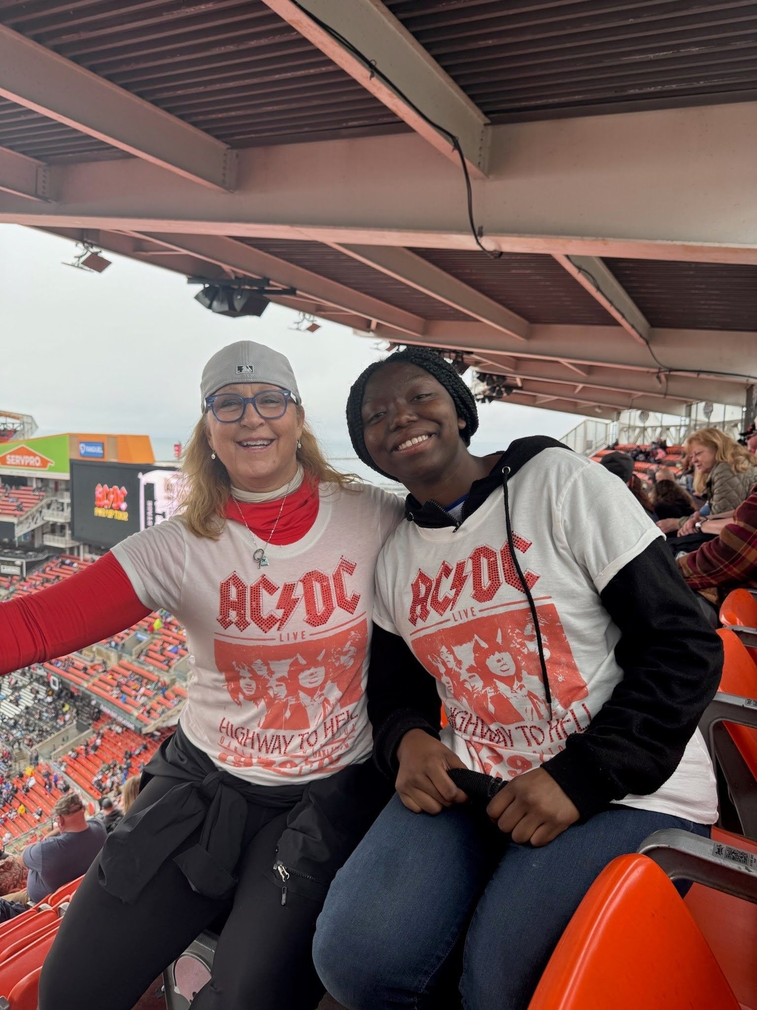 Big Sister Kelly with her Little Sister Aria in matching AC/DC shirts smiling in a Cleveland stadium seating area