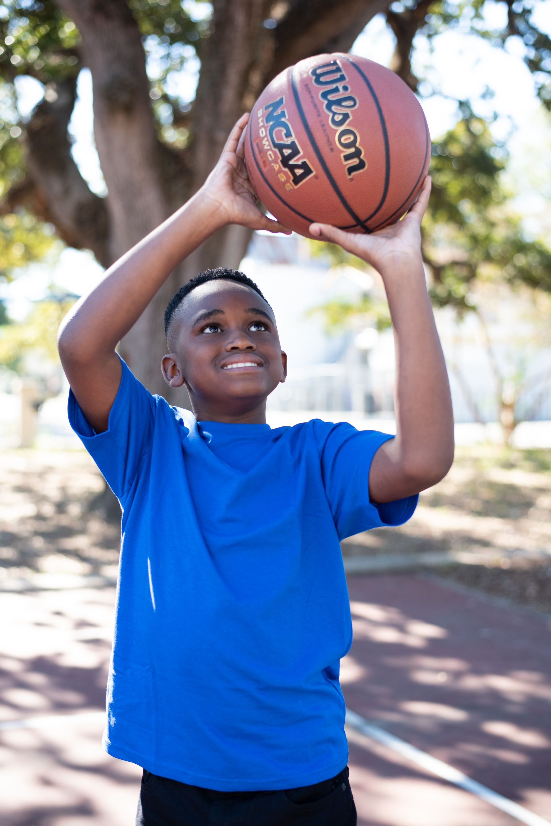 Little Brother in blue shirt holding a basketball overhead outdoors on a sunny day
