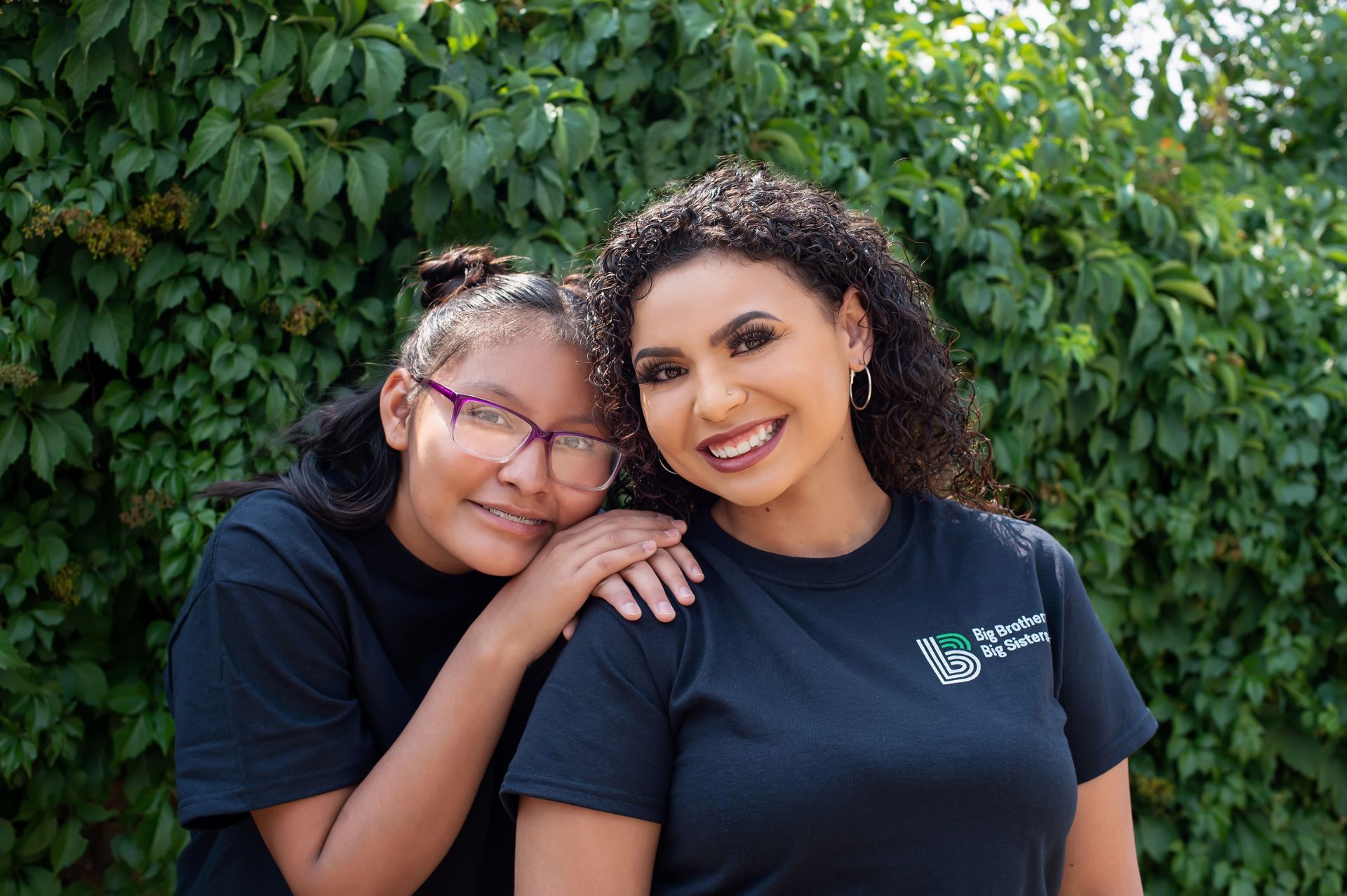Big and Little in black shirts posing in front of a leafy green wall