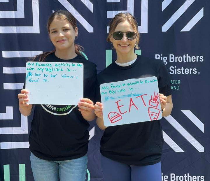 A Big and Little holding handwritten signs in front of a Big Brothers Big Sisters banner.