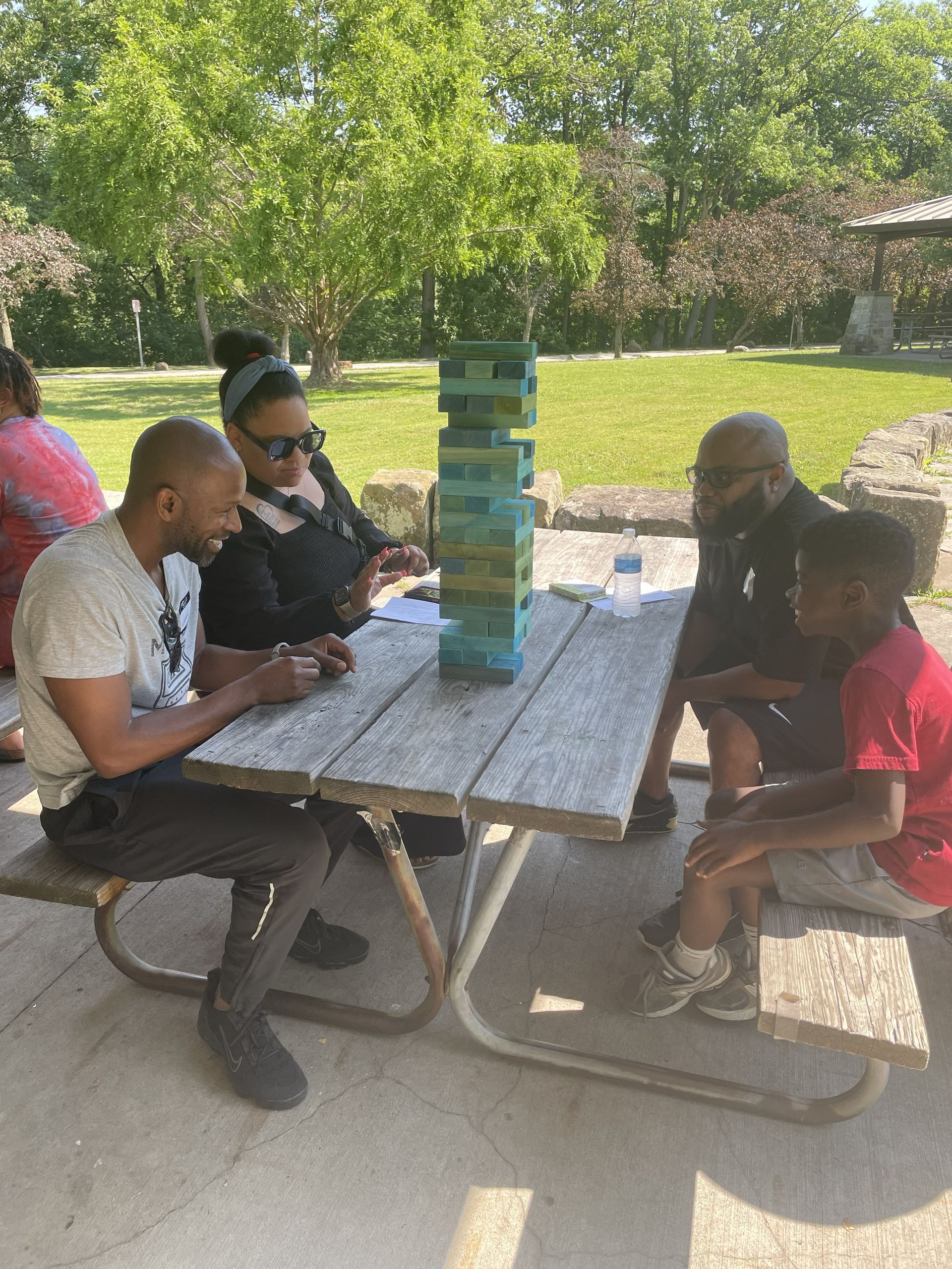 Big for a Day volunteers at a picnic table outdoors, playing a stacked block game in a sunny park with a Little at a Big for a Day event.