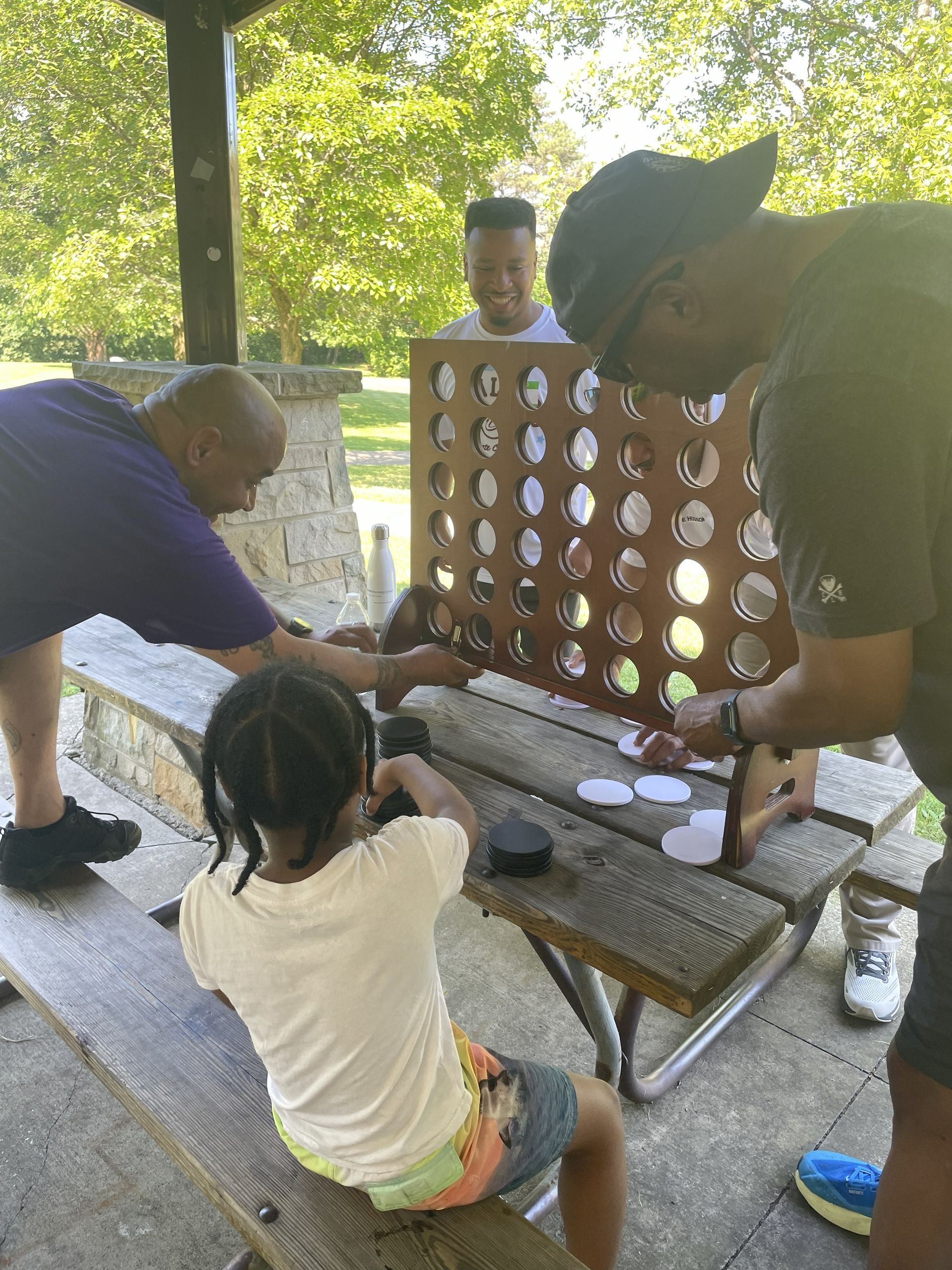 Big for a Day volunteers playing Connect Four at a picnic table in a park, with a child seated in the foreground.
