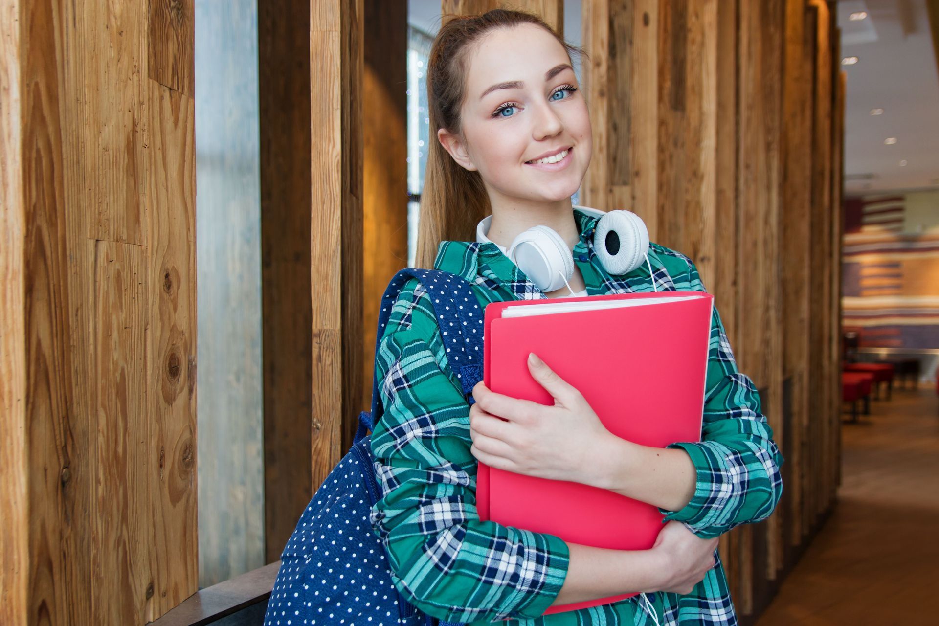 Student smiling in a hallway, holding a red binder and blue backpack over her shoulder.
