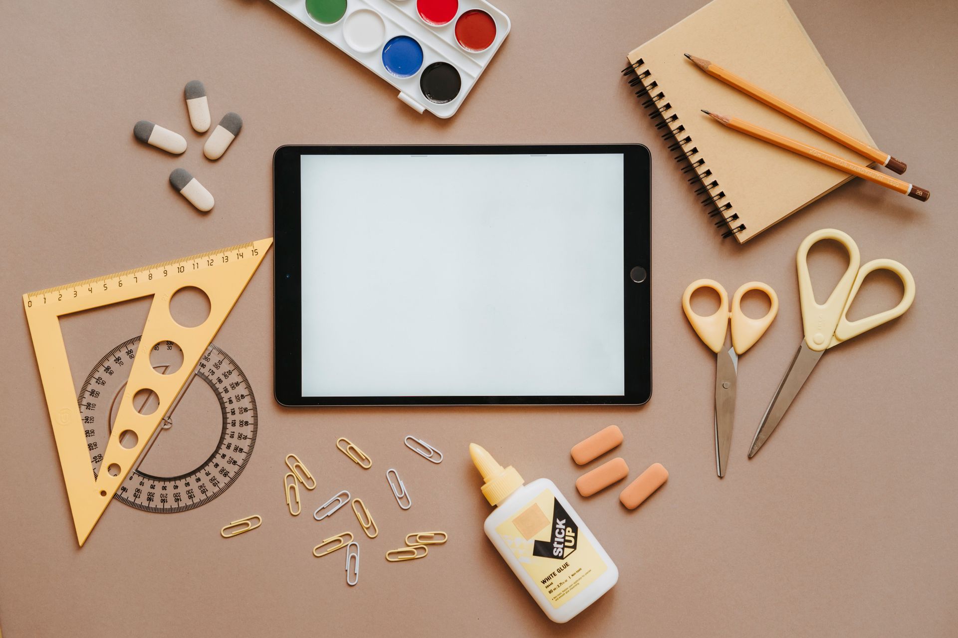 Craft supplies around a blank tablet on a beige desk, including paint, pencils, scissors, rulers, glue, and clips