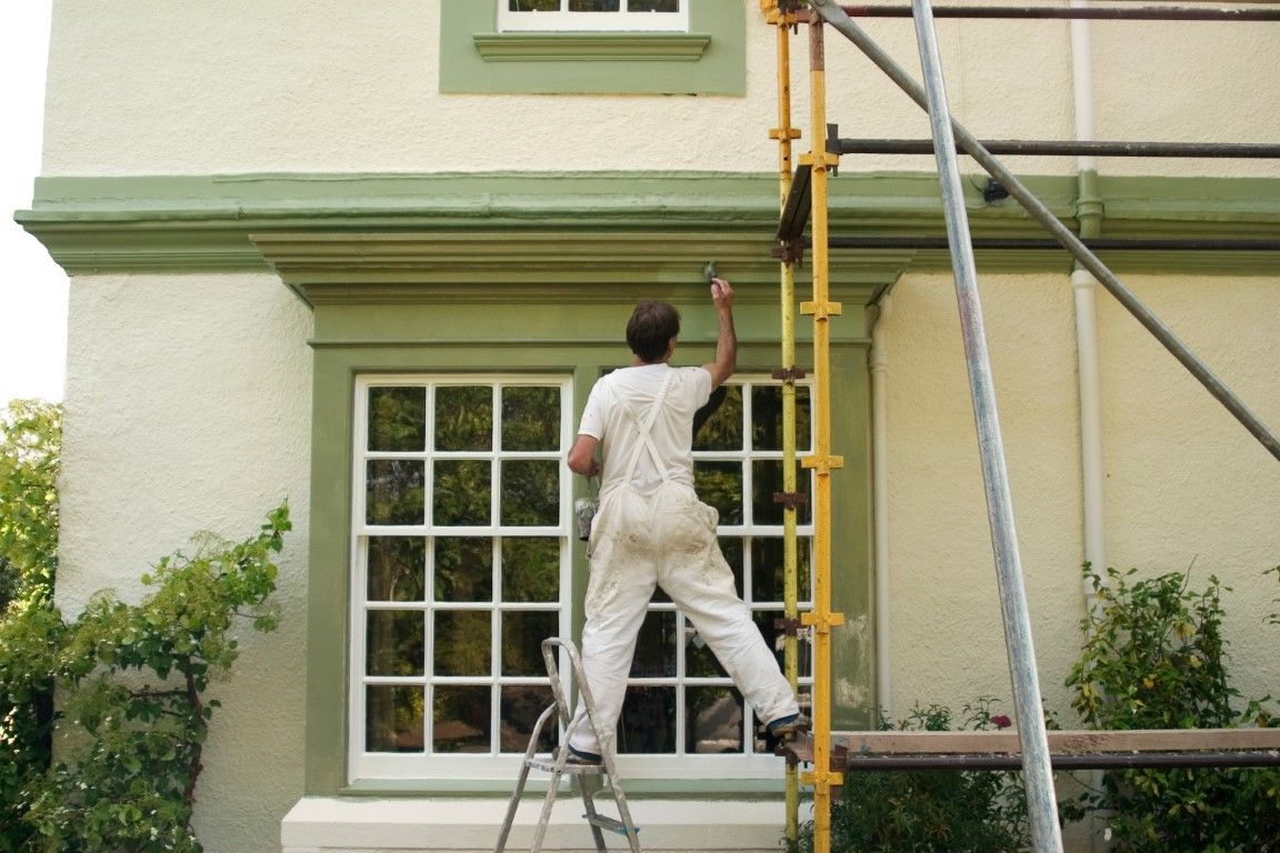 a man standing on a ladder outside, painting window trim green with a brush