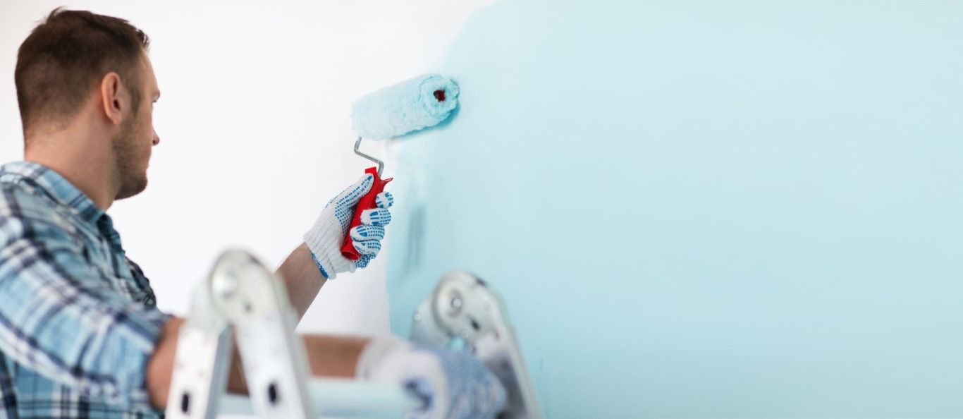 a close up of a man holding a paint roller painting a wall light blue at the top of a ladder