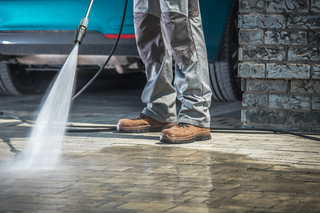 Person wearing work boots power washing a concrete surface with a car and brick wall in the background.