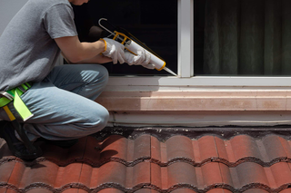 Person applying sealant around a window, kneeling on a red-tiled roof.