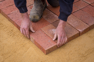 Person laying red brick pavers on a yellow sand base.