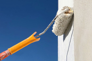 Paint roller applying white paint to a white wall against a blue sky.