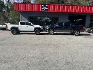A white and a dark-colored pickup truck parked side-by-side in front of a building with a red corrugated metal roof.
