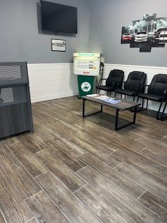 A waiting room with wood-look tile floors, three black chairs, a coffee table, a retro gas pump, and a TV mounted on a wall.
