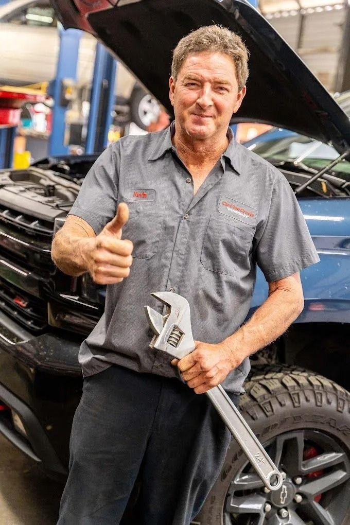 A mechanic wearing a grey uniform stands in a garage by a car with its hood open, smiling and giving a thumbs up.
