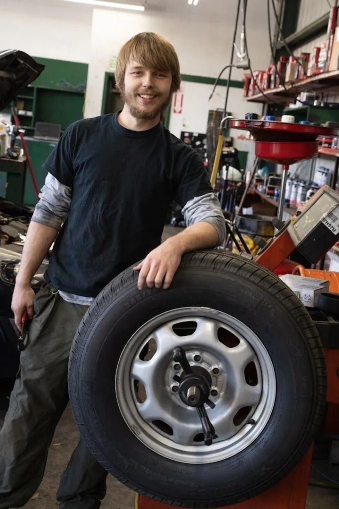 A smiling mechanic with a beard wearing a black t-shirt, resting an arm on a tire mounted on a wheel balancer in a shop.