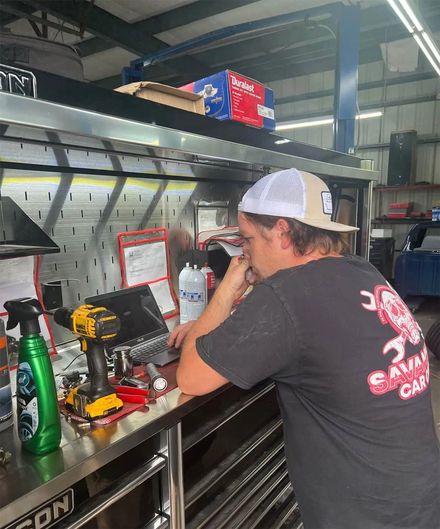 A person wearing a cap and t-shirt works on a laptop at a stainless steel workbench in an auto repair shop.