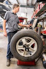 A technician in a gray work shirt operating a digital tire balancing machine inside an auto repair shop.