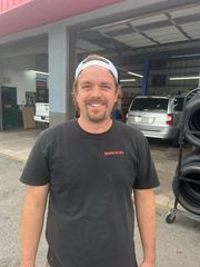 Smiling person wearing a white baseball cap and a black t-shirt standing in an auto repair shop with tires in the background.
