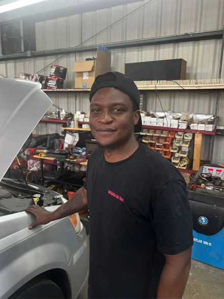 A person wearing a black t-shirt and cap stands beside a car with its hood open in an auto repair shop.