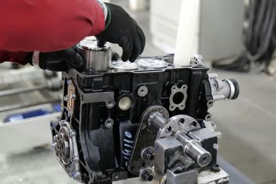 A technician in a red uniform and black gloves assembles a black engine block in a workshop.
