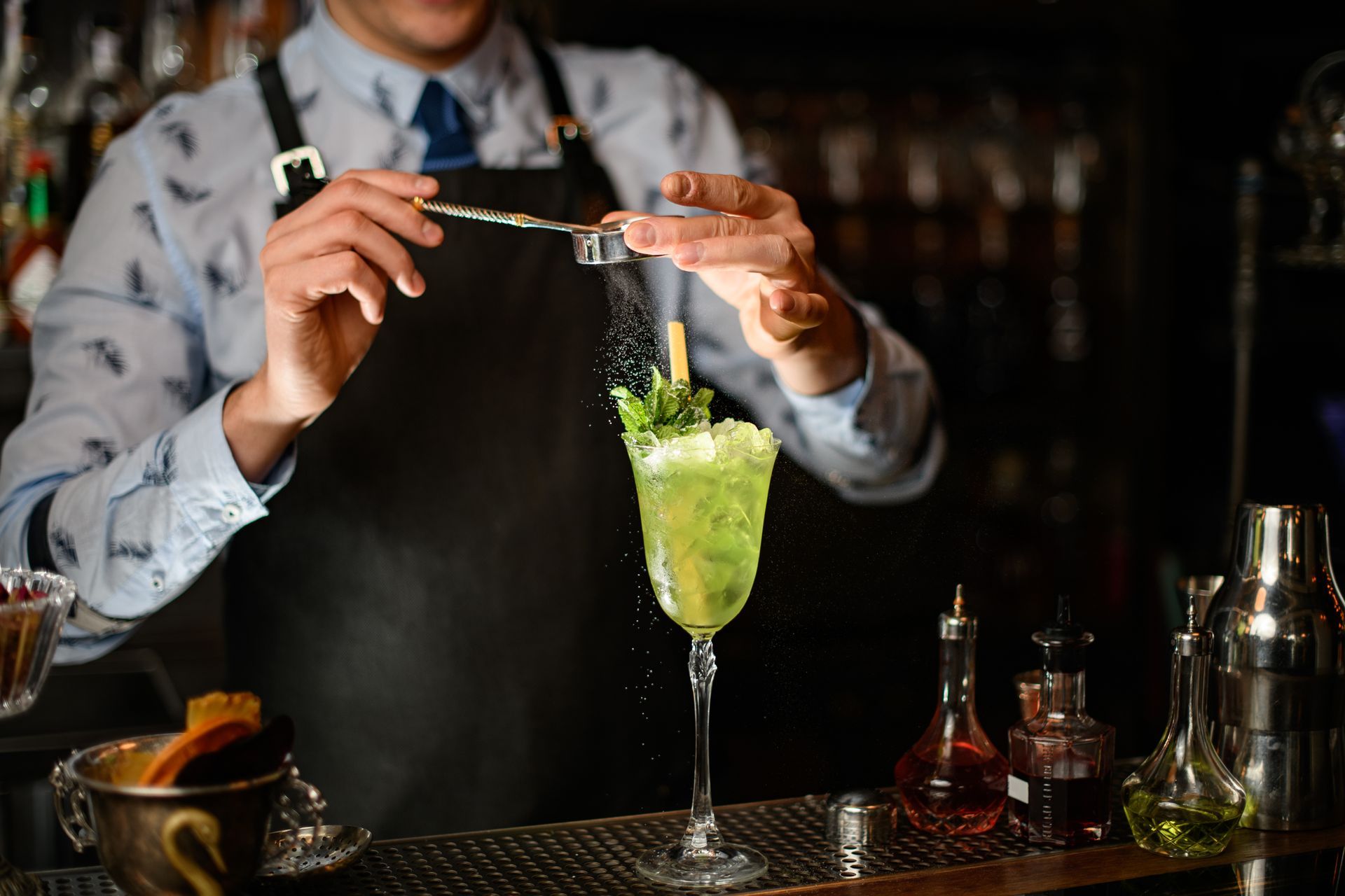 Bartender preparing a green cocktail with tongs at a bar counter.