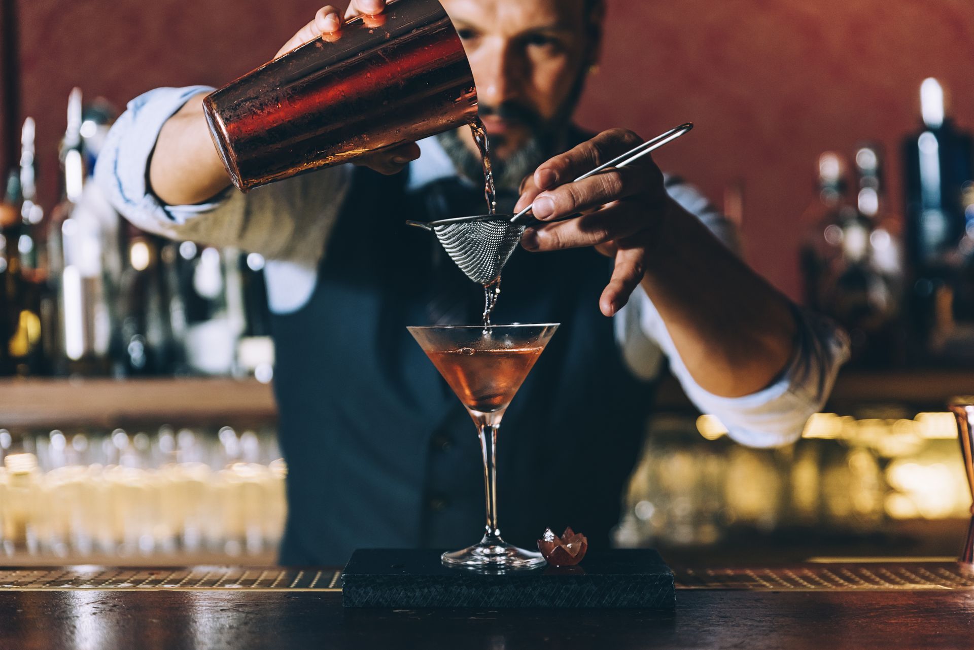 Bartender pouring a martini into a chilled glass at a bar, with a cocktail shaker in hand