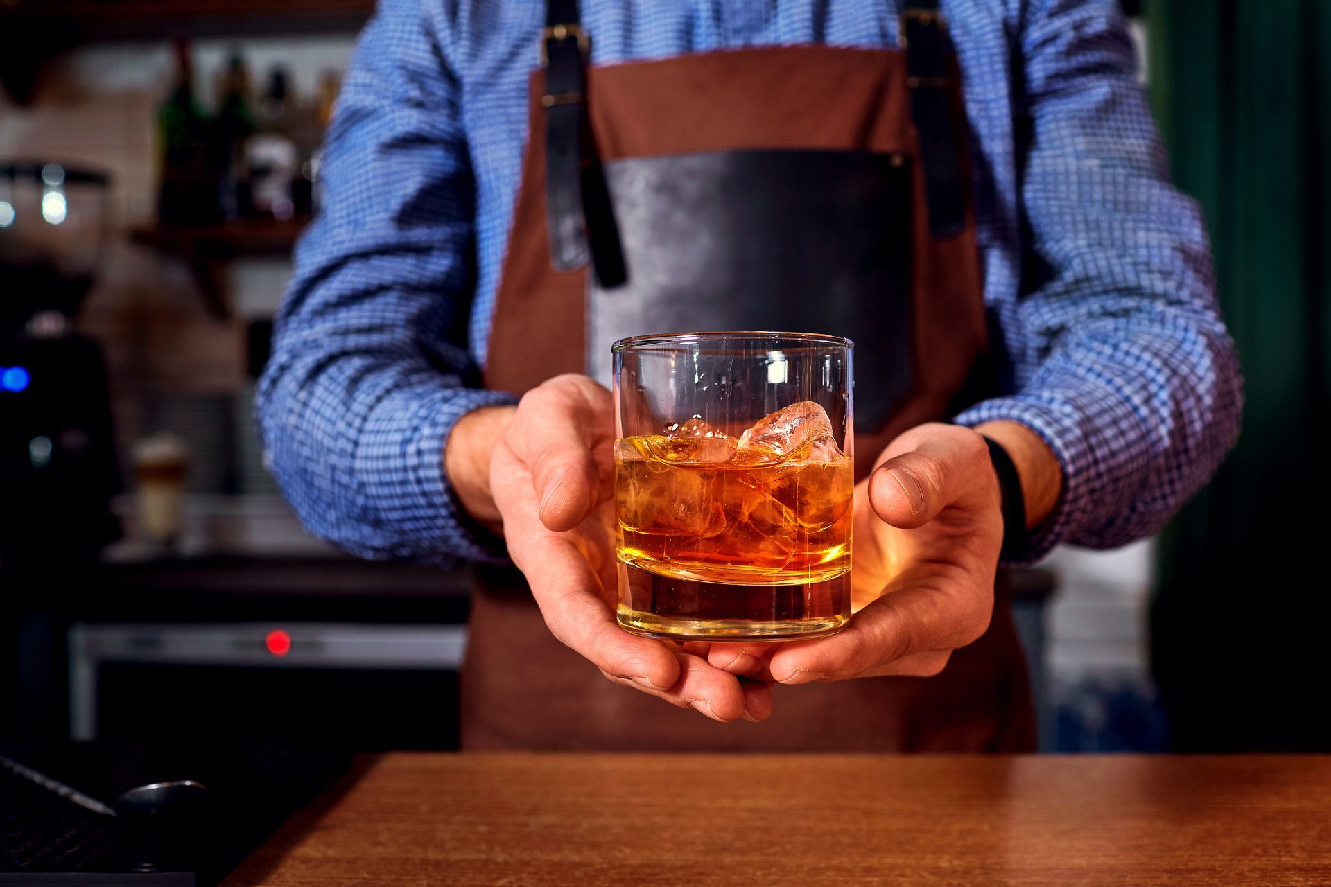 Bartender holding a whiskey glass with ice behind a wooden bar counter
