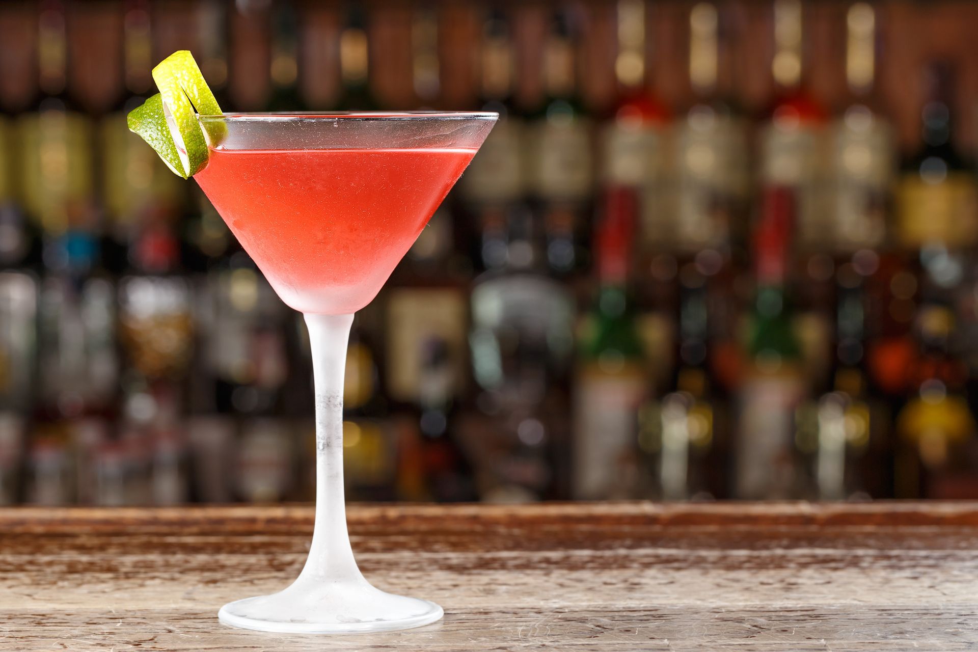 Pink cocktail in a martini glass with a lime wedge, set on a bar counter in front of blurred bottles
