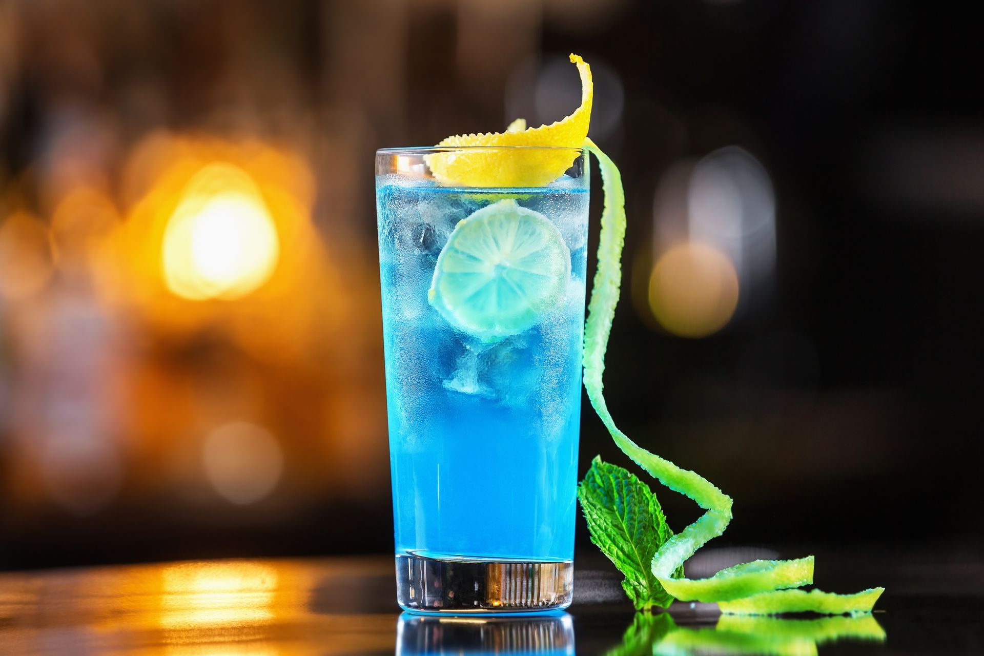 Blue cocktail with ice, lemon garnish, and a green garnish on a bar with blurred warm lights