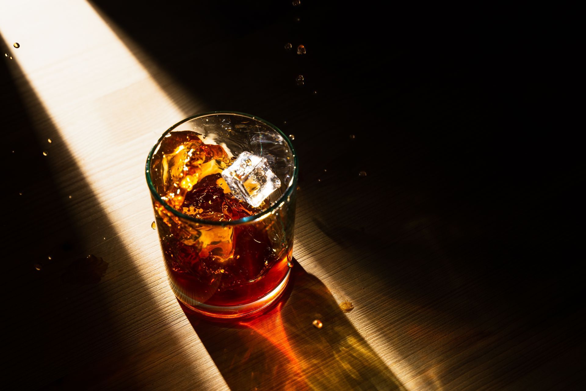 Glass of amber whiskey with ice on a sunlit wooden table in a dark room