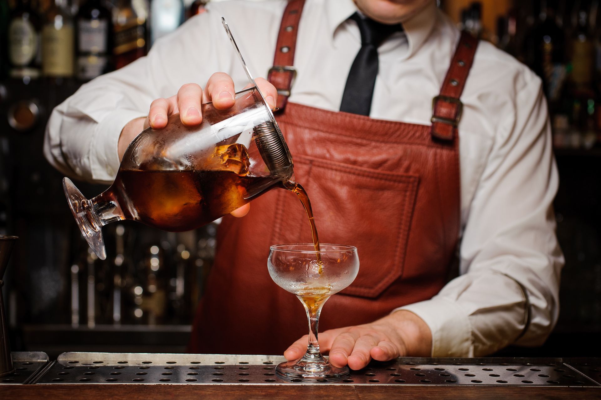 Bartender in white shirt and brown apron pouring amber drink into a cocktail glass behind a bar.