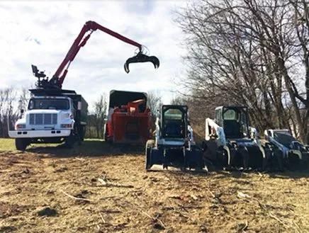 White crane truck loading machinery in a muddy field beside several blue tractors and bare trees