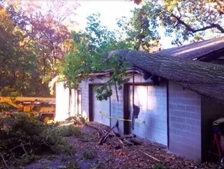 Rubble and fallen tree branches beside a damaged white house with a partially collapsed roof