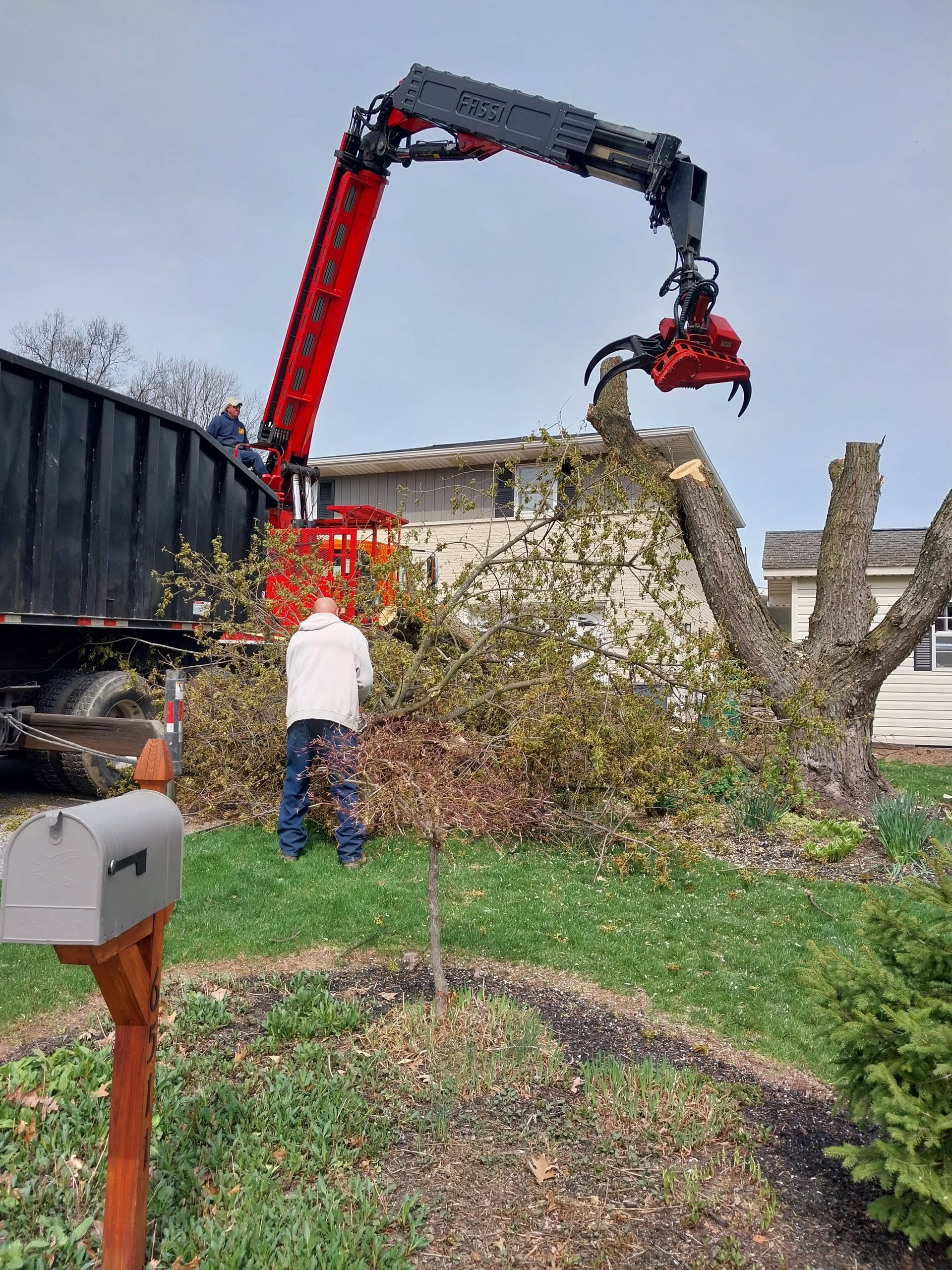 Red crane arm lifting debris from a pile beside a house, with a person standing nearby in a yard.