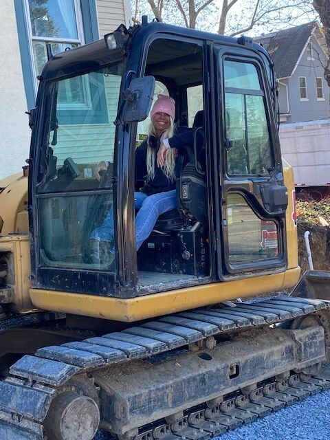 Person sitting inside a blue-and-yellow excavator cab outdoors