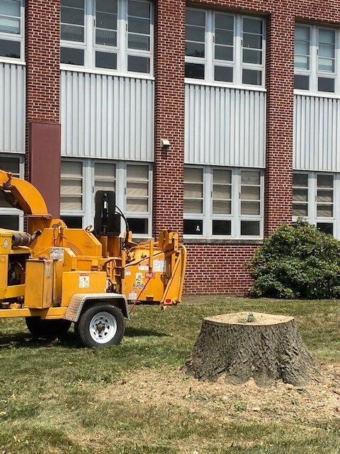 Yellow wood chipper beside a brick building, with a tree stump in the grass foreground.