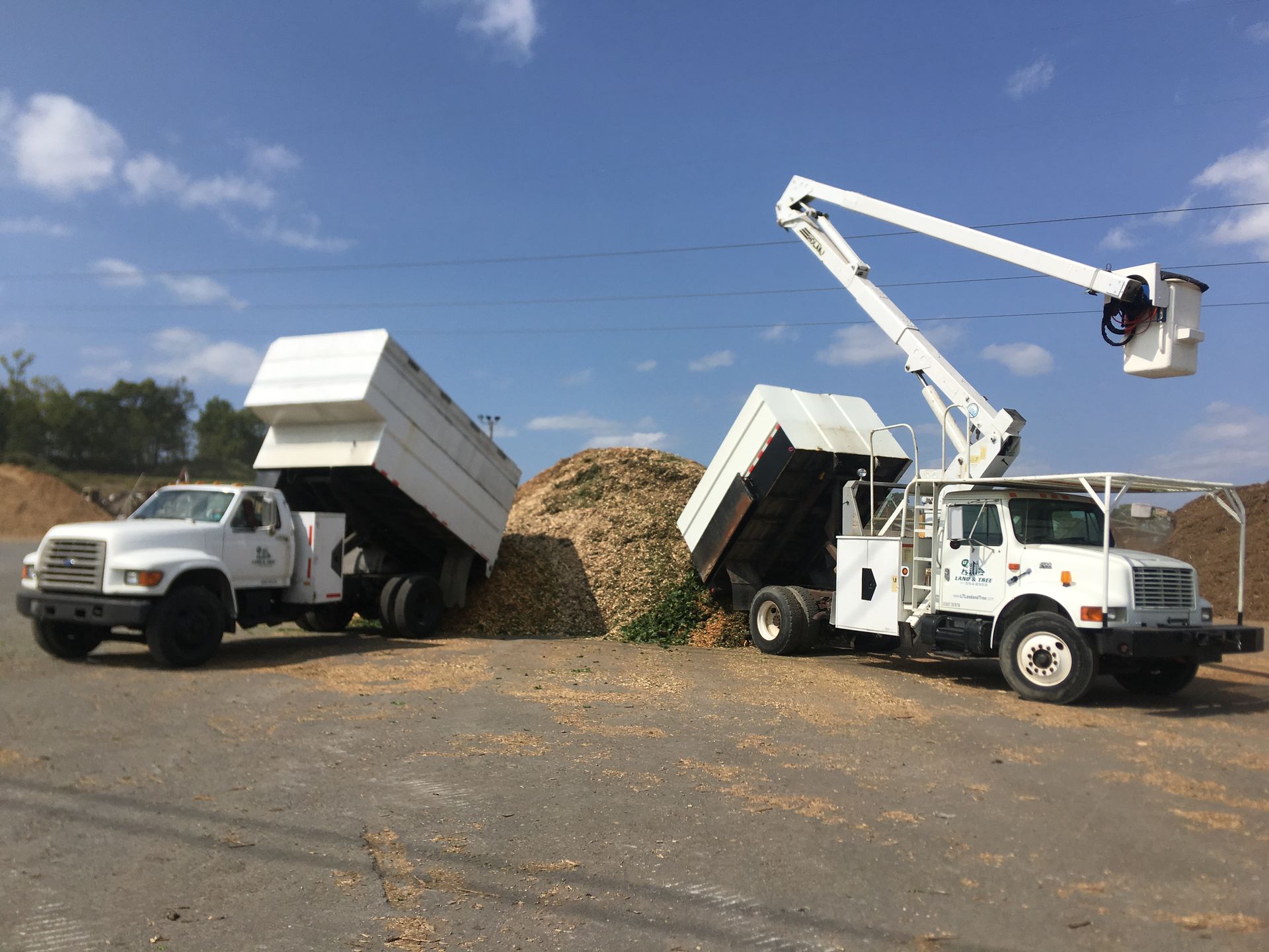 Two white dump trucks unloading a pile of wood chips in a yard, one with a boom lift raised overhead