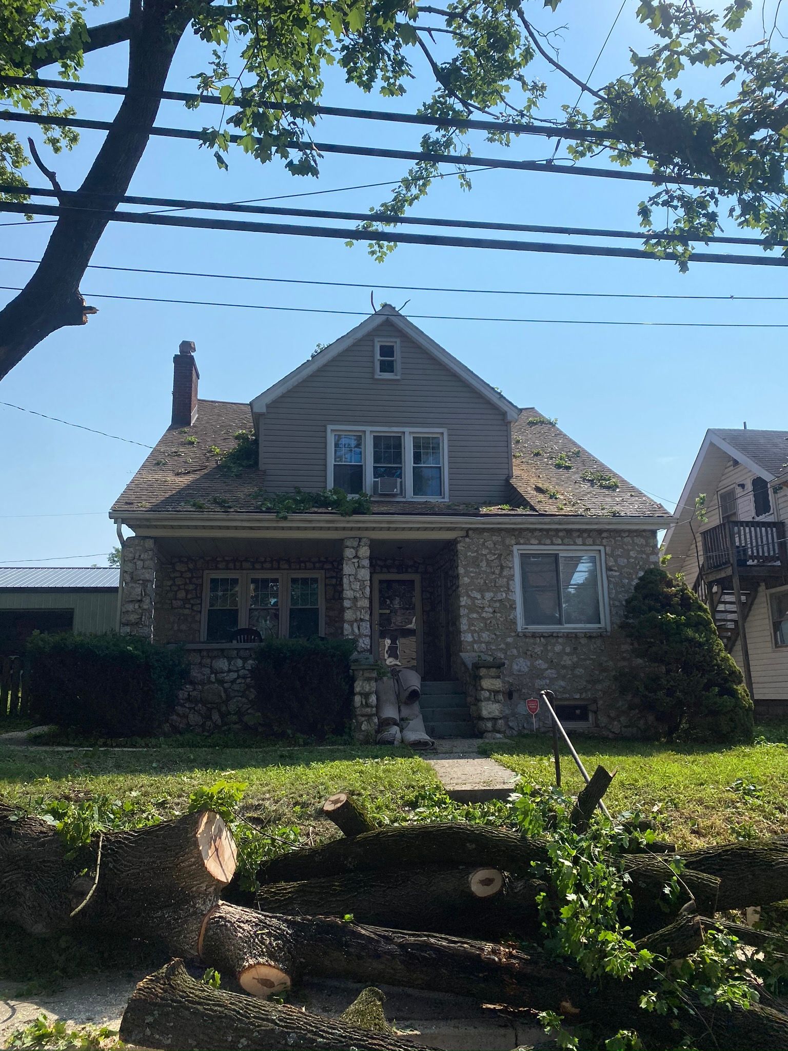 Small stone house with a front porch and dormer windows, seen from a grassy yard with tree branches overhead.