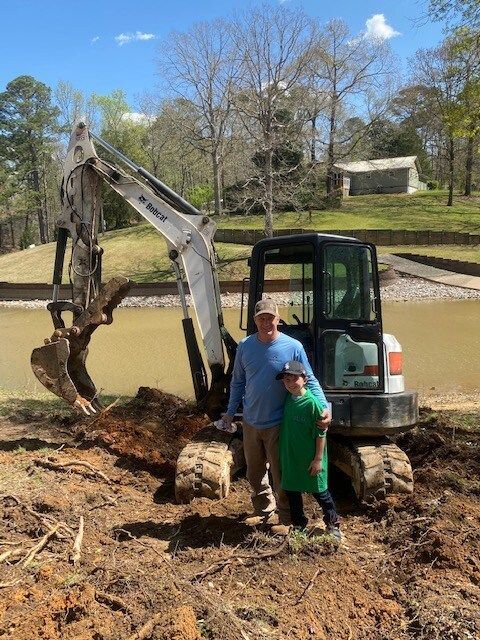 Two people stand beside a small excavator with its bucket in dirt at a sunny outdoor site.
