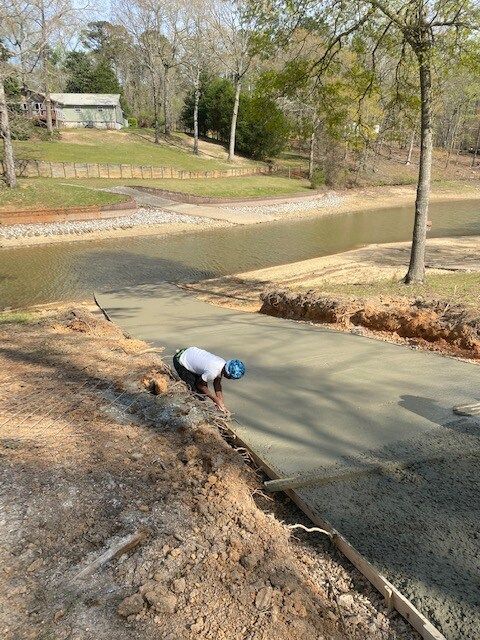 Worker crouching by a roadside ditch, spreading gravel along a newly paved path in a wooded area.