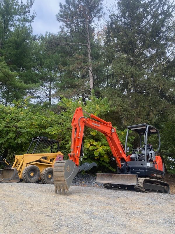Orange mini excavator beside a yellow dump truck on gravel with trees in the background