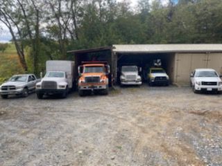 Five parked pickup trucks and vans under a shed in a gravel lot, with trees in the background