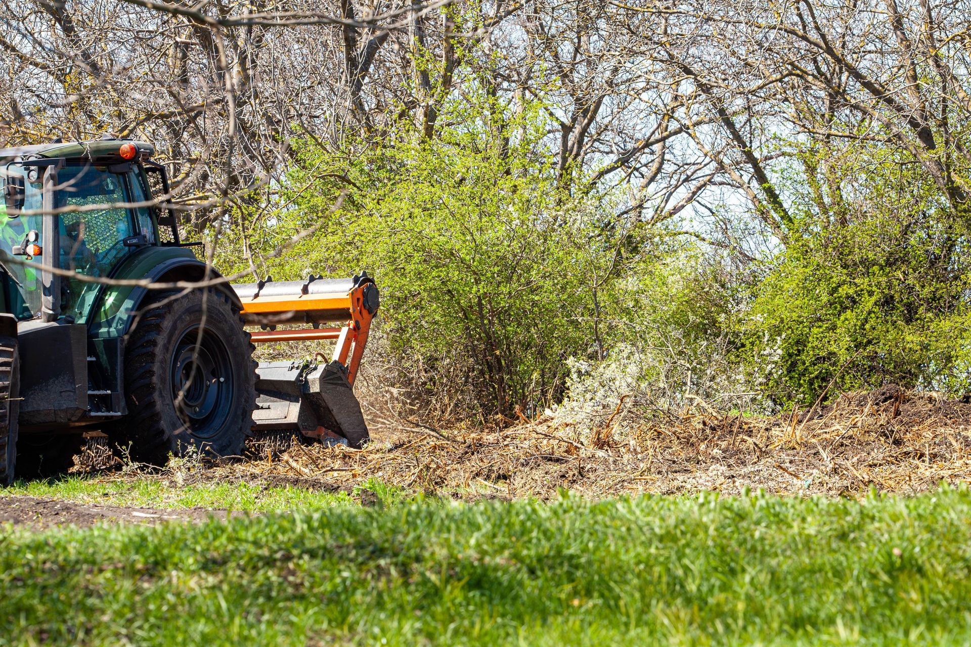 Tractor with mower clearing brush beside a grassy field and trees.