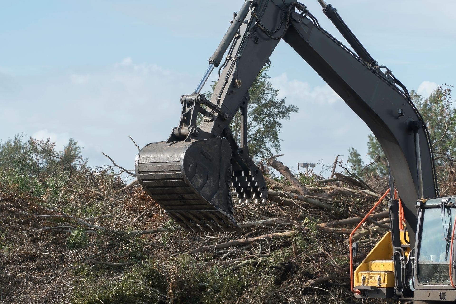 Excavator claw digging through a pile of branches and debris at a construction site.