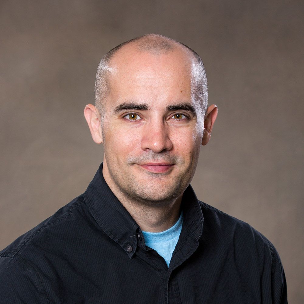 A bald man wearing a black shirt and a blue shirt is smiling for the camera.