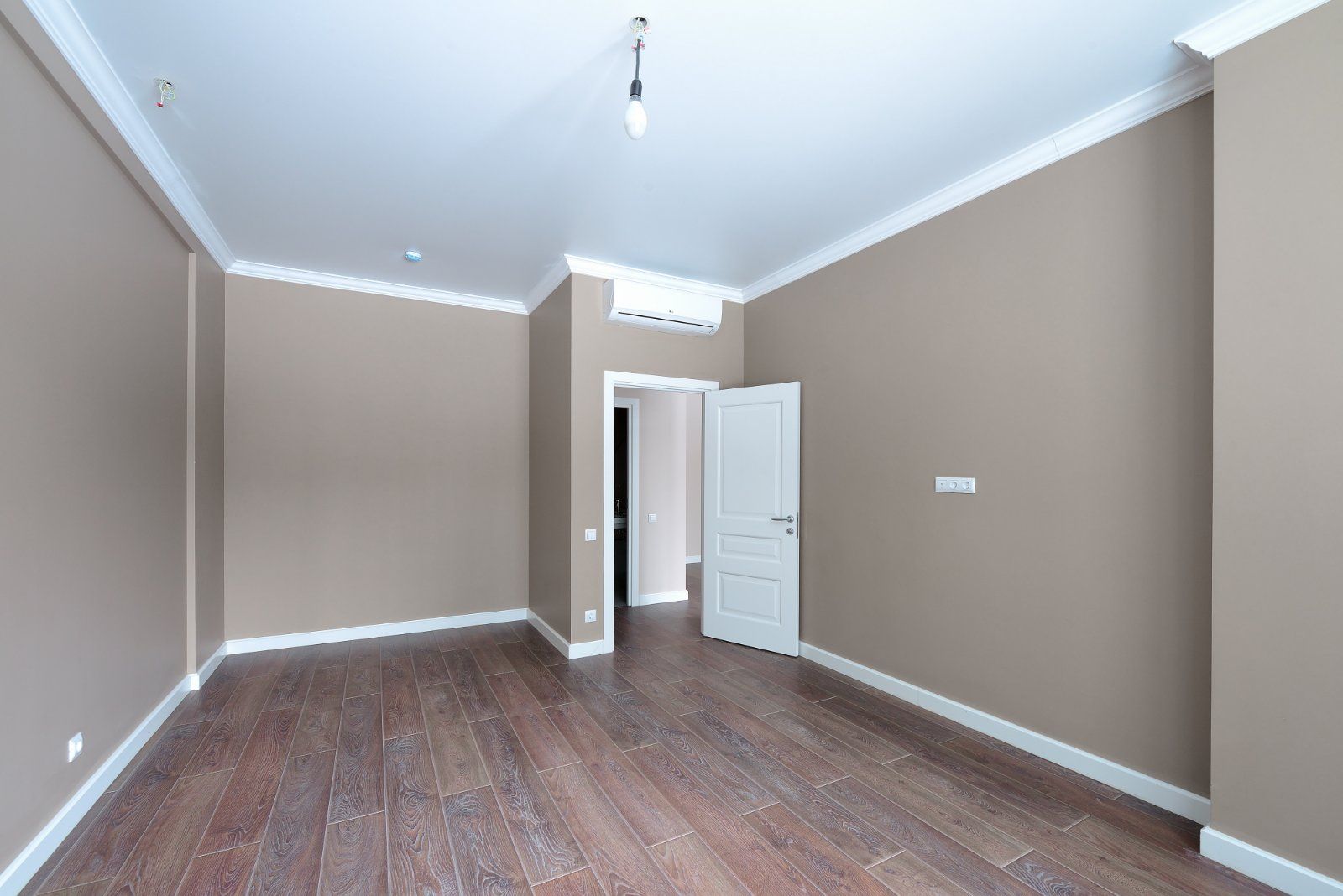 Empty room with brown walls, white trim, and hardwood floors. Open door leads to another dark room.