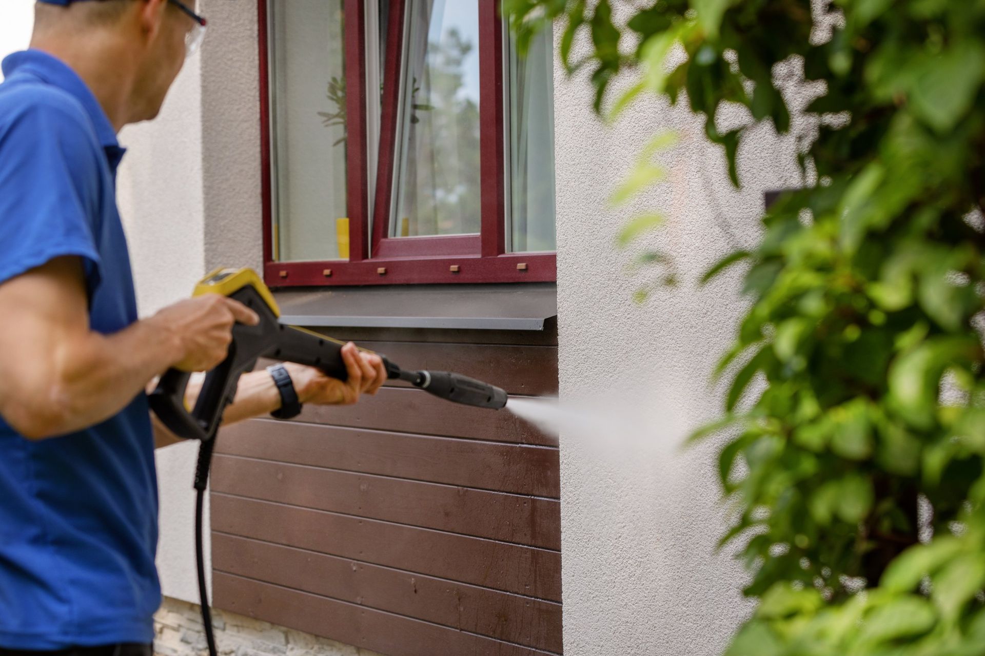 Person power washing exterior wall with pressure washer, near a window and greenery.
