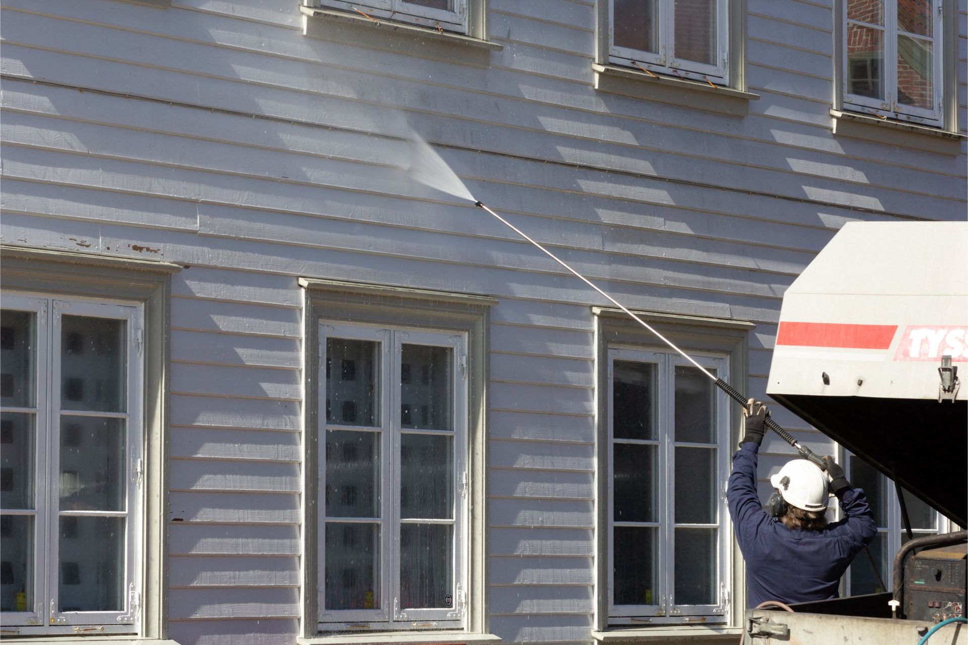 Man in safety gear power washing the exterior of a white wooden building with multiple windows.