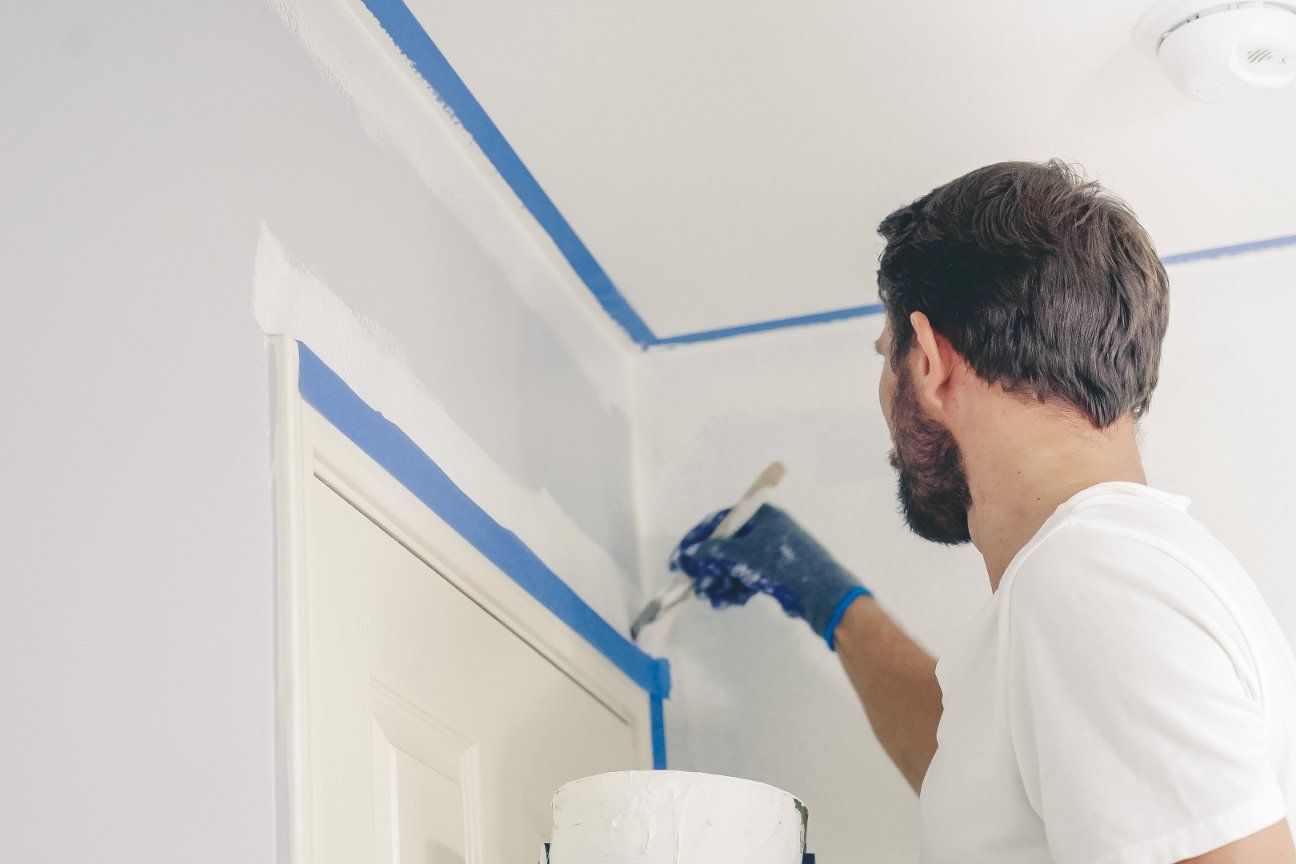 Man painting a wall blue, wearing gloves, with blue painter's tape on trim.