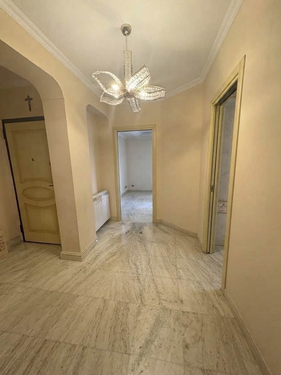 Corridoio vuoto color beige con pavimento in marmo, lampadario e porte che conducono alle stanze adiacenti.